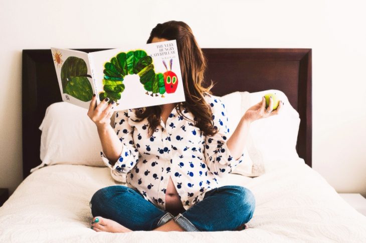 Sesión de fotos de maternidad chica leyendo un libro a su bebé mientras está sentada en la cama 