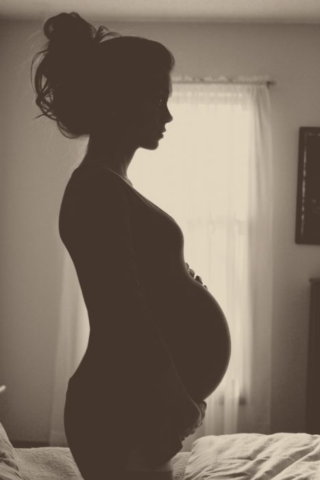 Sesión de fotos de maternidad chica parada junto a una ventana con un contra luz