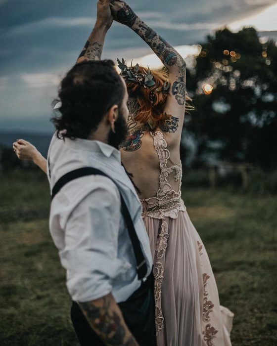 Novios bailando en su boda vikinga, esposa con vestido rosa de encaje sin mangas y tatuajes en los brazos, novio con barba, camisa blanca y tirantes