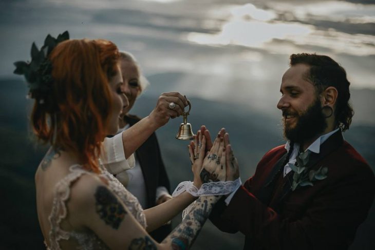 Pareja de prometidos se casa en boda nórdica, esposa con vestido de encaje rosa pálido y esposo con traje rojo vino, tomándose las manos