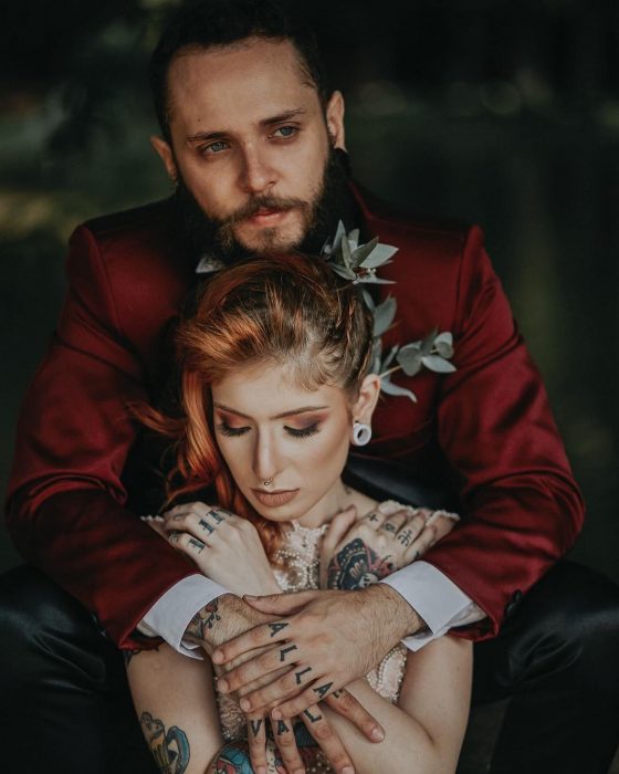 Pareja celebra boda vikinga, novio con traje rojo vino con negro abrazando a esposa con vestido de encaje y cabello pelirrojo