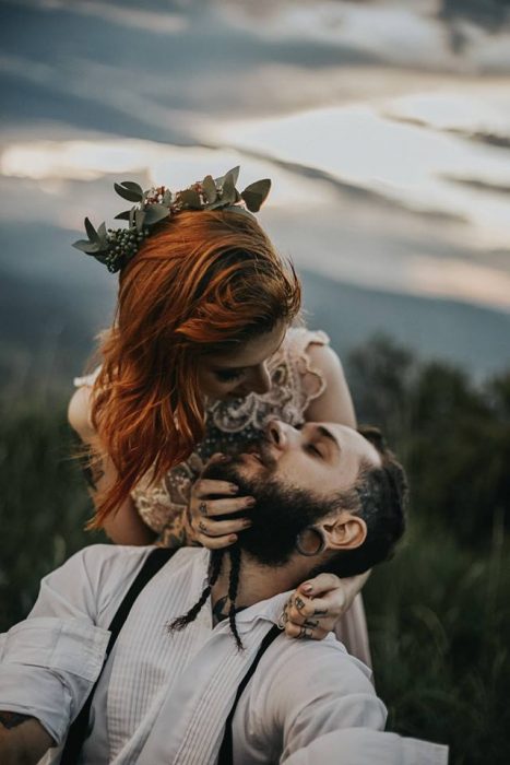 Boda vikinga, novia pelirroja con tocado de hojas y vestido de encaje mira y abraza a novio con barba y expansiones en las orejas