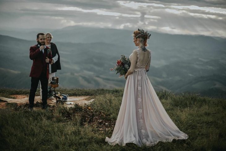 Pareja de novios con tatuajes celebra una boda vikinga al aire libre, esposa con vestido de encaje rosa y esposo con traje rojo