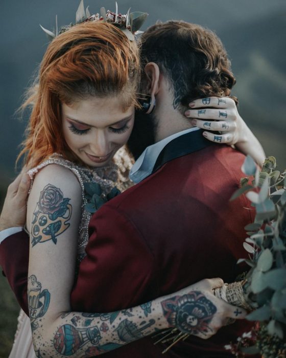 Novios se casan en boda vikinga, mujer pelirroja y con tatuajes abraza a novio con traje rojo vino