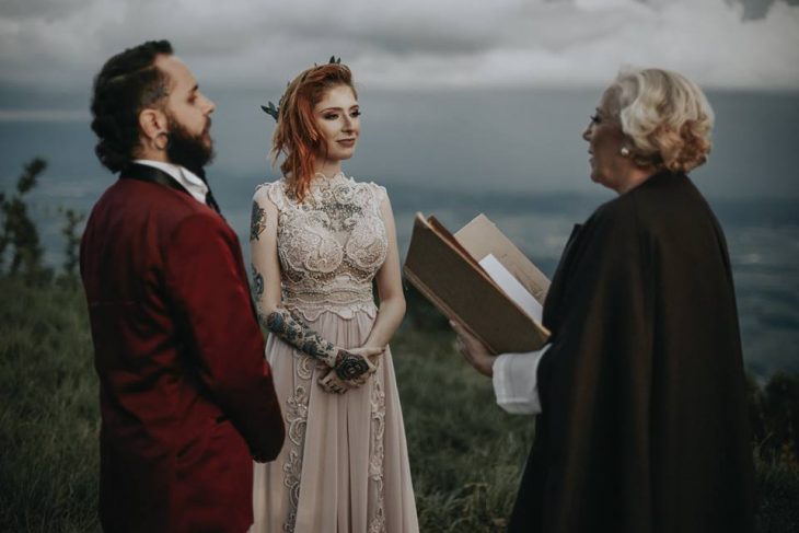 Pareja de prometidos se casa en boda nórdica, esposa con vestido de encaje rosa pálido y esposo con traje rojo vino, novios parados frente a jueza