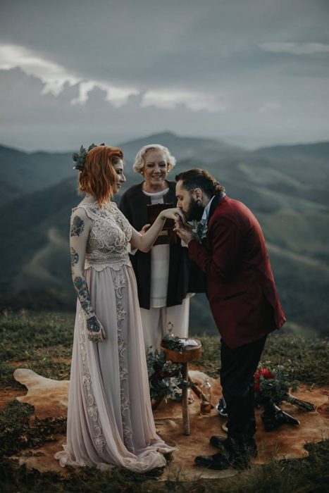 Pareja de novios con tatuajes celebra una boda nórdica al aire libre, novio con traje guinda besa la mano de su prometida con vestido largo vintage