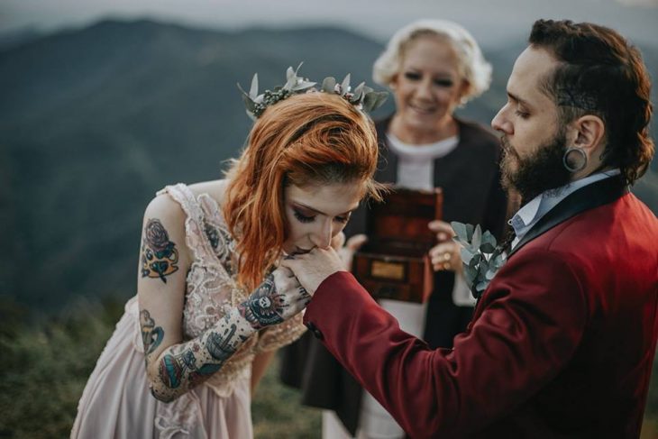 Pareja de novios con tatuajes celebra una boda nórdica al aire libre, novia de cabello rojo besa la mano de su novio con saco guinda