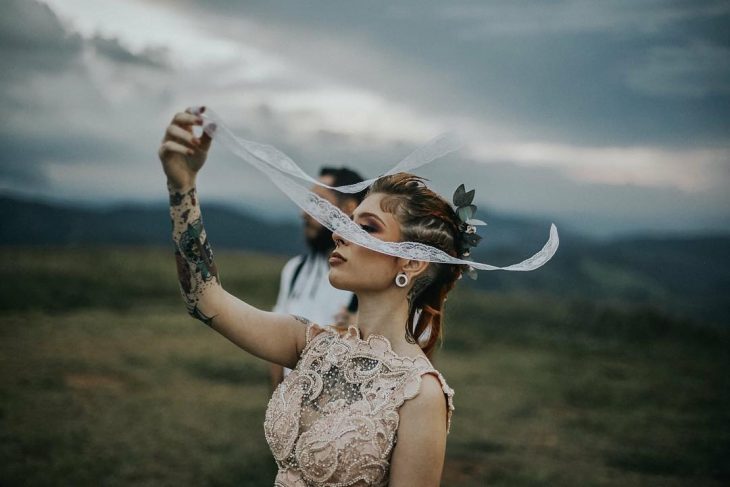 Pareja de novios celebra una boda nórdica al aire libre, mujer con tatuajes y expansiones con vestido de encaje con perlas sostiene listón de encaje blanco