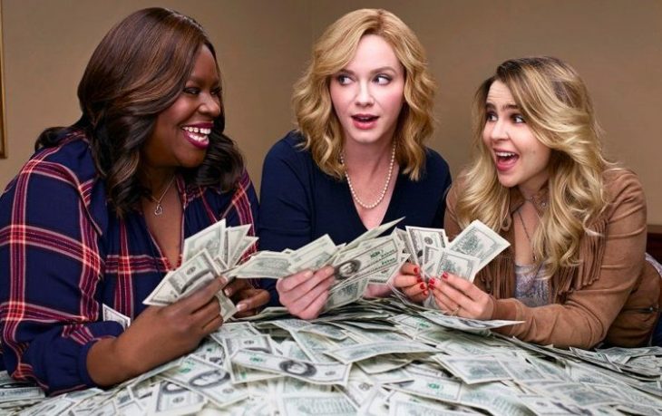 Mujeres reunidas alrededor de una mesa sosteniendo dinero entre sus manos, mirándose sorprendidamente entre sí, escena de la serie Chicas buenas