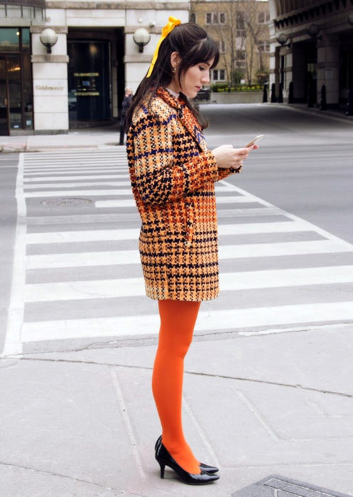 Outfits con medias de colores; chica con medias anaranjadas y con saco de cuadros, mirando su celular en la calle