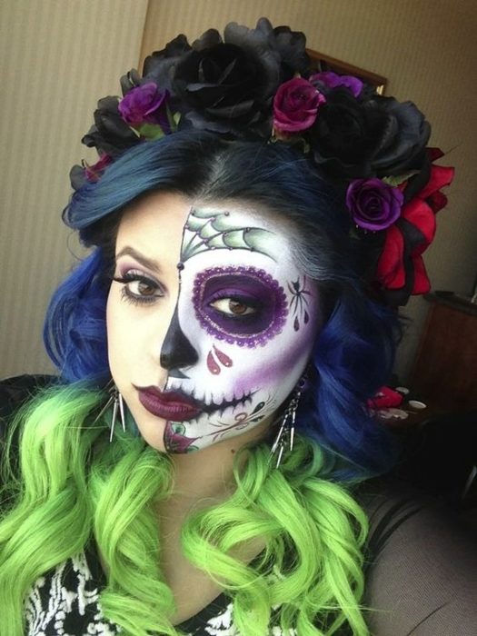 Maquillaje de catrina sobre la mitad del rostro, en colores negro, blanco y morado, con corona de flores del mismo color