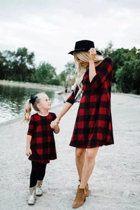 Madre e hija tomadas de la mano cerca de un lago las dos con vestido rojo como cuadros negros, la madre usa botines cafés y sombrero negro con el cabello largo, rubio y ondulado y la niña usa mallones negros con botas plateadas con coleta alta de lado y un moño negro grande