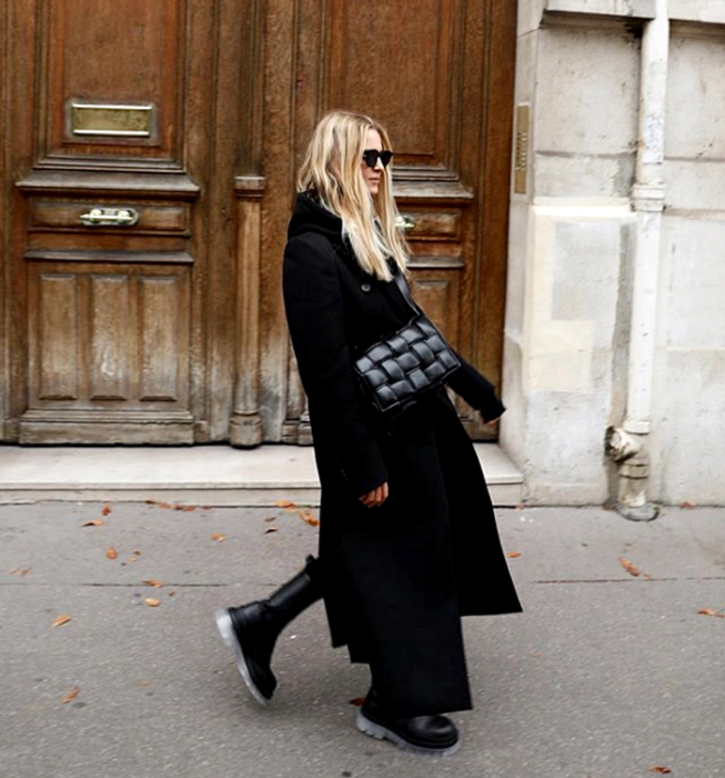 chica rubia con lentes de sol, usando un top negro, leggings negros, abrigo largo negro, bolso de mano negro de piel y botas de suela gruesa negras