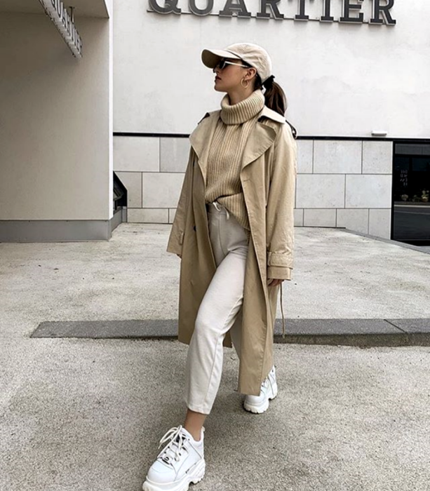 chica de cabello castaño usando una gorra beige, suéter tejido beige de cuello alto, abrigo largo beige, pantalones de vestir beige, tenis blancos deportivos